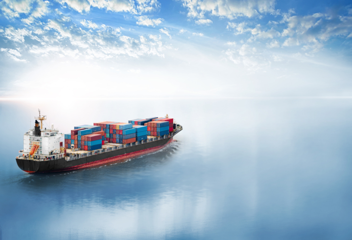 Cargo container ship sailing across calm blue ocean waters under a bright sky with scattered clouds.