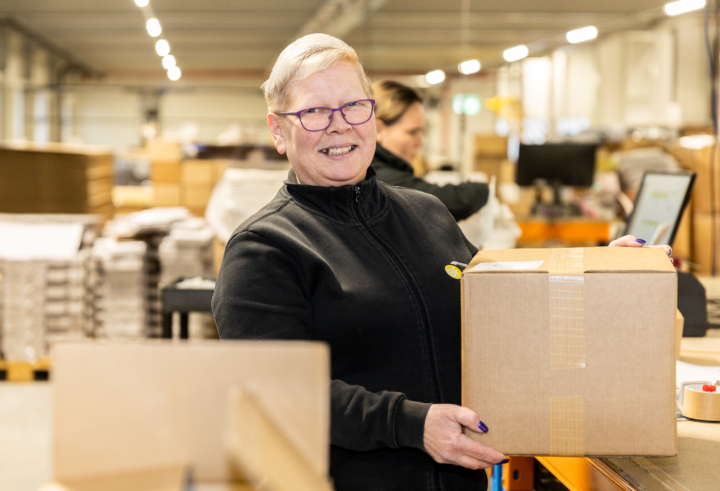 Lachende magazijnmedewerker met een kartonnen doos in een druk distributiecentrum.