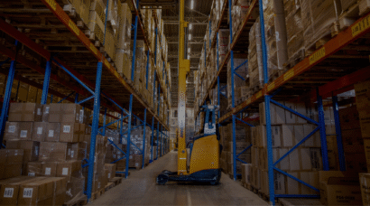 Smallegangentruck rijdt door een gangpad in een hoogbouwmagazijn, omringd door hoge palletstellingen vol kartonnen dozen.