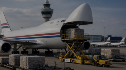 Photo of a white cargo aircraft with red and blue striping unloading freight at Schiphol Airport, with cargo pallets, ground handling equipment, and airport buildings in the background.
