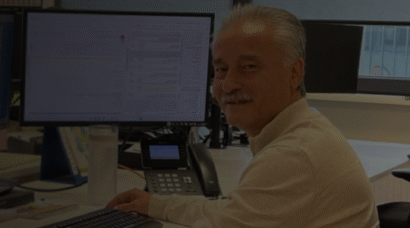 Smiling man at a desk in an office, looking toward the camera, with a computer monitor and phone on the workstation.